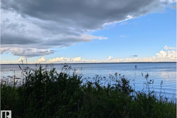 View across a wide, calm lake with tall grasses in the foreground and a dynamic, cloudy sky above. - 334 Crystal Springs Dr, Rural Wetaskiwin County
