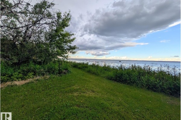 Expansive lake view from a manicured lawn bordered by trees and vegetation, with a cloudy sky overhead. - 334 Crystal Springs Dr, Rural Wetaskiwin County