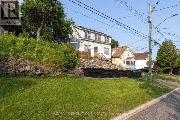 Side view of a white multi-story house with a rocky embankment and green grass in front. - 334 Pearl Street, Thunder Bay