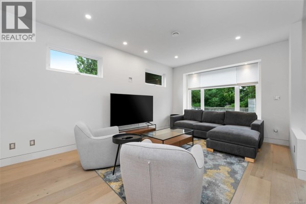 Living room with gray sectional sofa, two armchairs, a glass coffee table, and large windows with green outdoor views. - 3341 Skinner Rd, Duncan