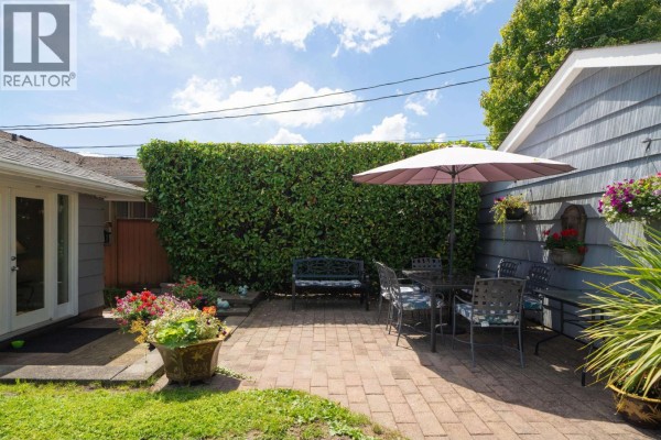 Paved backyard patio with a table, chairs, umbrella, and surrounded by green hedges. - 335 Churchill Avenue, New Westminster