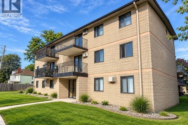 Rear exterior of the apartment building with a paved parking lot and trees. - 3355 Sandwich Street Unit# 304, Windsor