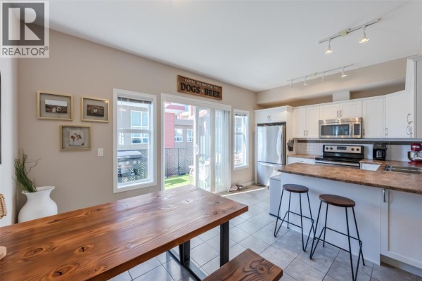 Kitchen and dining area with white cabinets, stainless steel appliances, wooden table, and backyard view. - 3363 Wilson Street Unit# 125, Penticton