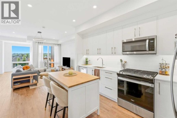 Open concept white kitchen with island, stainless steel appliances and living room. - 338, 110 18a Street Nw, Calgary