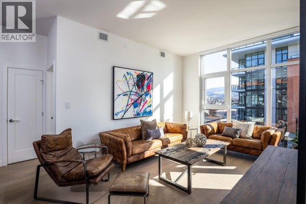 Living room with brown leather sofas, abstract art, and large windows with city views. - 3389 Lakeshore Road Unit# 413, Kelowna