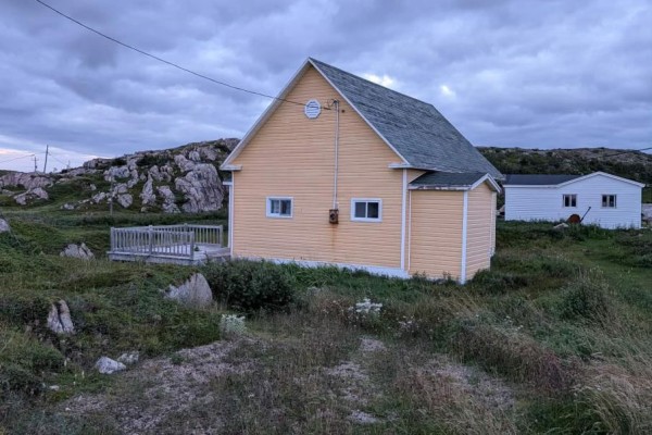 Side view of a yellow house with a gabled roof and light-colored siding in a rural setting. - 339 Main Road, Fogo Island