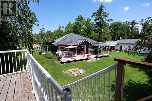 View from a deck looking towards the rear of a grey bungalow with a large lawn and trees. - 34 Coldstream Road, Kawartha Lakes