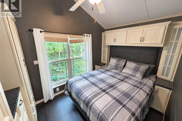 Mobile home bedroom with a queen-size bed, plaid bedding, built-in cabinets, and a window overlooking trees. - 34 Temagami Trail, Wasaga Beach