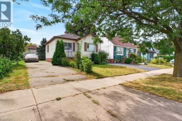 Side view of a detached bungalow with white siding, showing driveway and front yard with mature trees. - 340 Barton Street, Stoney Creek