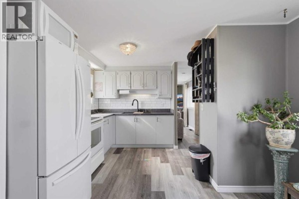 View from dining area into the white kitchen, showing grey cabinets, white appliances, and grey walls. - 341, 5344 76 Street, Red Deer