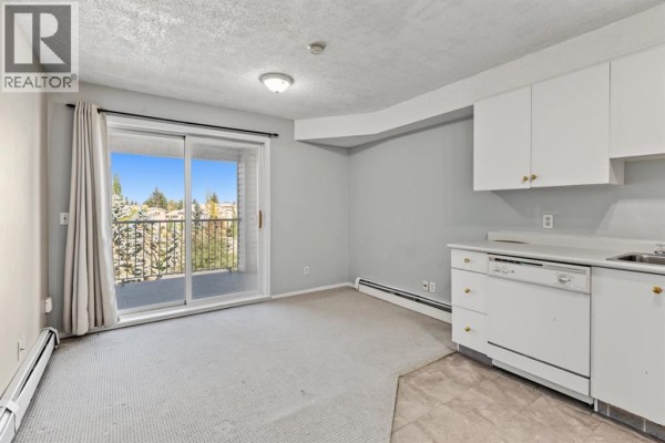Living area with beige carpet, sliding glass door to balcony, and white kitchen cabinets. - 3415, 1620 70 Street Se, Calgary