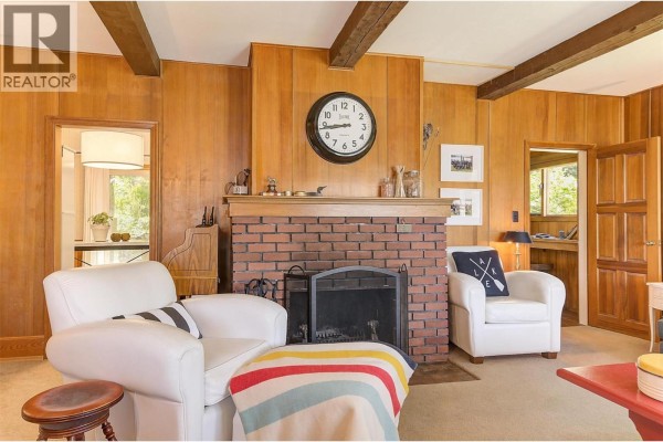 Living room with wood-paneled walls, brick fireplace, white chairs, and a striped blanket. - 3415-3421 Princeton Summerland Road, Princeton