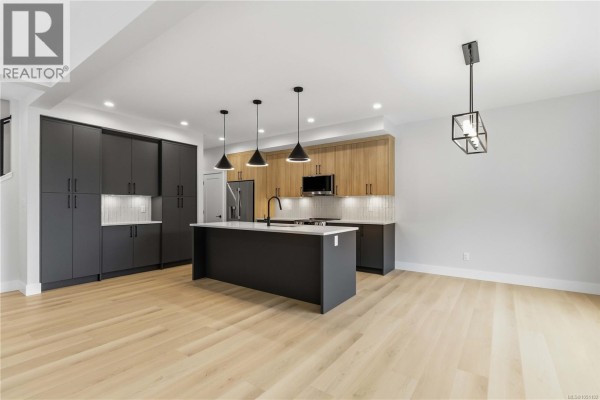 Open-concept living space with a modern kitchen featuring dark grey cabinets, wood accents, and an island. - 3462 Merlot Terr, Langford