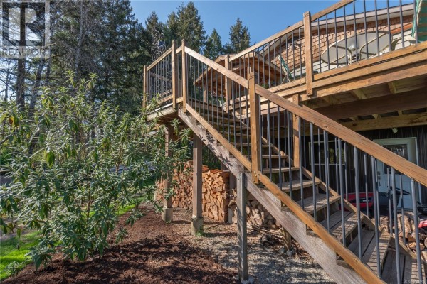 Close-up of the wooden deck stairs and railing with firewood stacked neatly on the ground below. - 3468 Cowichan Lake Rd, Duncan