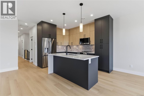 Kitchen view from the side showing stainless steel refrigerator, dark grey pantry, and light wood cabinetry. - 3482 Merlot Terr, Langford
