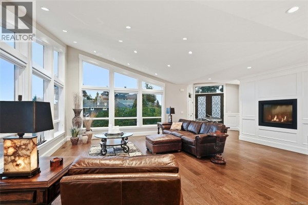 Living room with leather sectional, fireplace, and large windows overlooking greenery. - 3495 Simmons Pl, Nanoose Bay