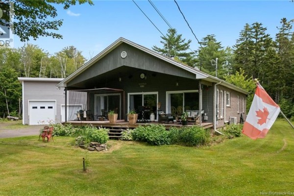 Side view of the grey house showing the entrance deck, Canadian flag, and surrounding trees. - 3515 Route 530, Grande Digue
