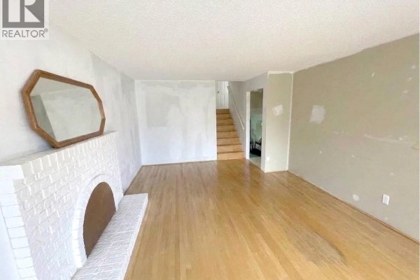 Living room with hardwood floors, white brick fireplace, and unfinished walls, showing stairs to an upper level. - 3520 Broadway Street, Richmond