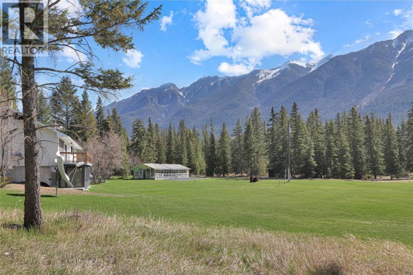 View of the house from a grassy field, with dense trees and snow-capped mountains beyond. - 3526 Island Pond Road, Skookumchuck