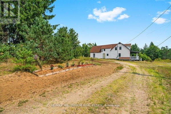White two-story house with a covered porch on a dirt driveway with a garden bed. - 3529 Highway 118 E, Bracebridge