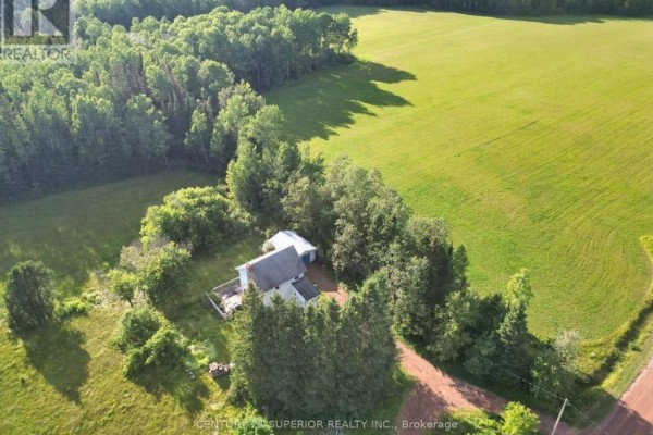 Aerial view showing the white farmhouse nestled amongst trees, bordering a vast, sunlit green field. - 353 Spring Creek Drive, Dorion