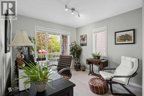 Home office with light gray walls, carpet flooring, a large window, and desk furniture. - 354 West Creek Springs, Chestermere