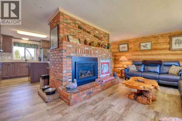 Living room featuring a large brick fireplace, log walls, leather sofas, and a view into the kitchen. - 3574 Spokin Lake Road, Williams Lake