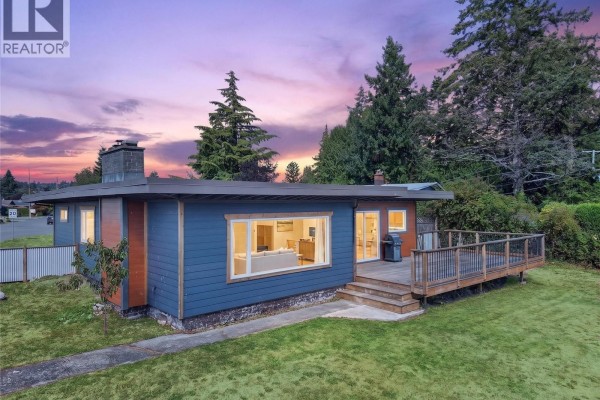 Dusk exterior shot of the ranch house with a deck and view into the lit living area. - 3584 Island Hwy, Campbell River
