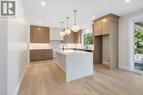 A modern kitchen with light wood cabinetry, a large marble island with pendant lights, and a window overlooking greenery. - 3586 Sutton Avenue, Windsor