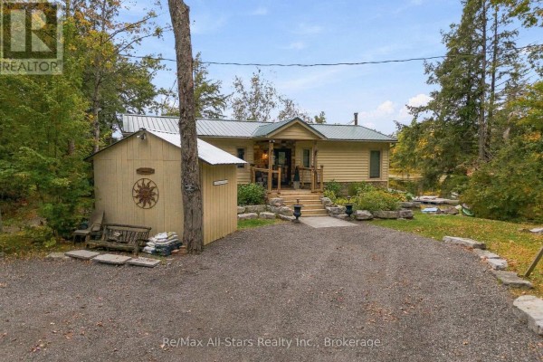 Front view of a tan, single-story house with a gravel driveway and surrounding trees. - 36 Jack Pine Way, Trent Lakes