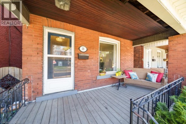 Close-up of the covered front porch with a wicker sofa, side table, and brick exterior. - 36 Sherman Avenue N, Hamilton
