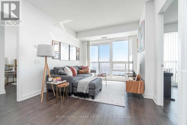 Living room with a grey sectional sofa, ottoman, rug, floor lamp, and large window with city view. - 3602 - 2212 Lake Shore Boulevard, Toronto
