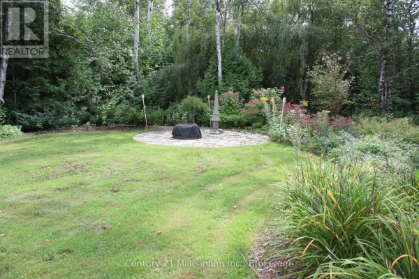 View of a circular stone patio in a grassy backyard surrounded by trees and landscaping. - 361 Tyendinaga Drive N, Saugeen Shores