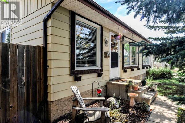 Close-up of the duplex entrance with beige siding, a wooden fence, and a small patio area with plants and a chair. - 3611b 66a Street, Camrose