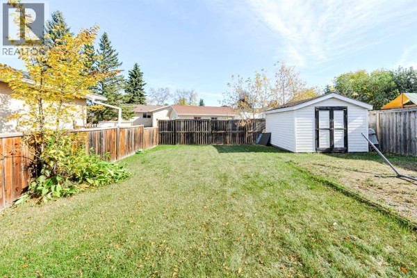View of a fenced backyard with a large green lawn, mature trees, and a white storage shed on the right. - 3611b 66a Street, Camrose