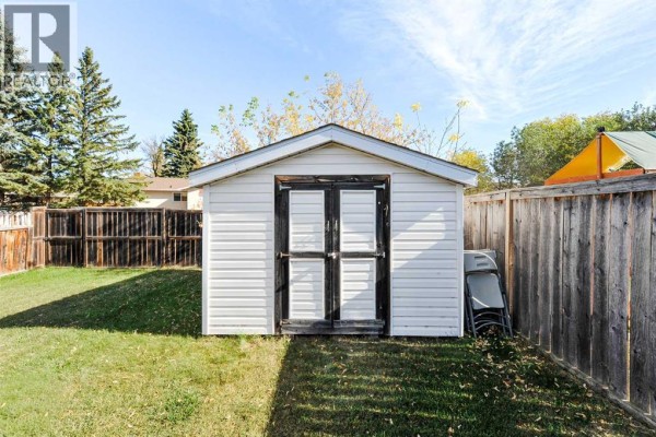 A white storage shed with double doors stands in a grassy backyard, partially shaded by trees and a wooden fence. - 3611b 66a Street, Camrose