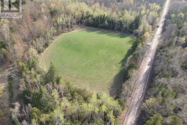 Aerial view of a cleared field surrounded by forest, with a paved road and a single vehicle visible on the right side. - 3643 10th Line East, Trent Hills