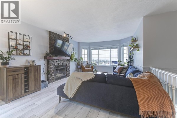Living room with a dark blue sectional sofa, a stone fireplace, and large bay windows. - 3656 Walnut Glen Drive, West Kelowna