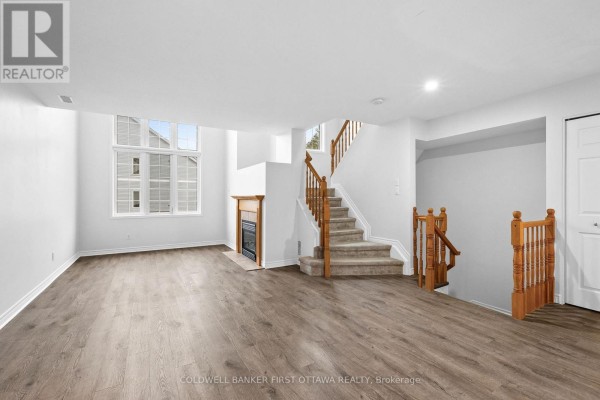 Living room with wood-look vinyl flooring, carpeted stairs, fireplace, and large windows overlooking the exterior. - 366 Wiffen Private, Ottawa