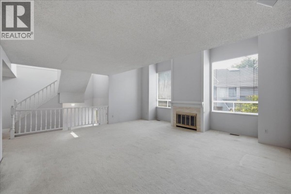 Spacious living room with a fireplace, beige carpeting, and large windows overlooking trees. - 3661 Handel Avenue, Vancouver