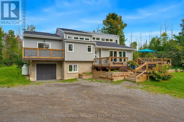 Front view of a grey house with decks, garage, and surrounding trees under a blue sky. - 368 Ramsay Concession 5a Road, Mississippi Mills