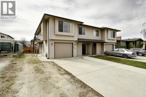 Exterior view of the beige duplex building with attached garages and driveway parking. - 369 Wallace Road, Kelowna