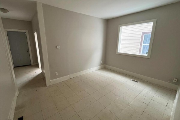 Interior room with tiled floor and light gray walls. A window looks out to a building. An entryway is visible on the left. - 370 Pritchard Avenue, Winnipeg