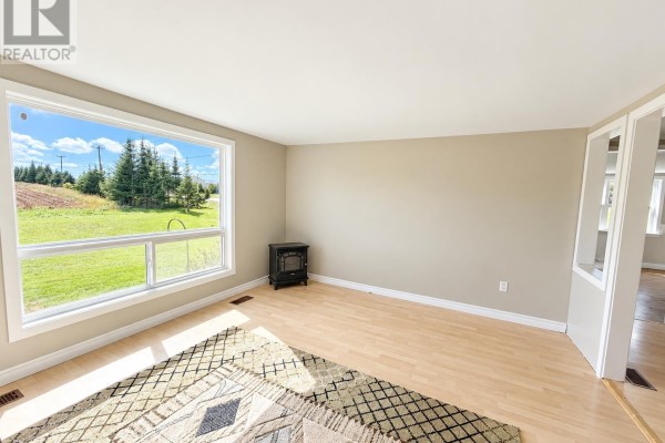 Living room with light wood floors, a large window, a small black wood stove, and a patterned rug. - 3702 North Carleton Road, Rte 10, Searletown