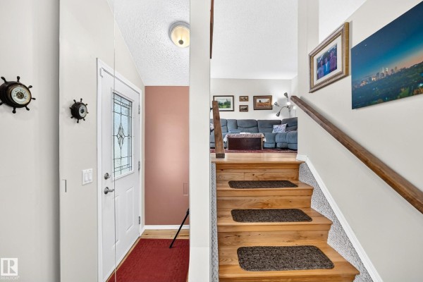 Interior view of a hallway with a white door, a staircase with wooden steps, and a view into the living room. - 371 Kirkpatrick Cr Nw, Edmonton