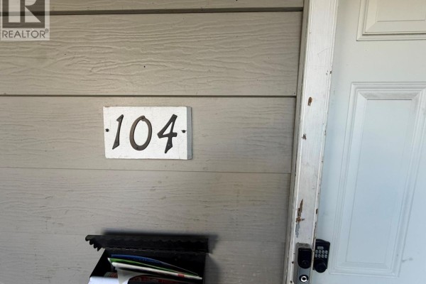 Close-up of unit number 104 mounted on beige siding next to a white door. - 3710 24 Avenue Unit# 104, Vernon