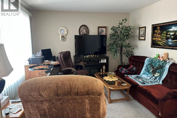 A living room with beige carpet, a red sofa, and a desk with a computer. - 3712 24 Avenue Unit# 105, Vernon