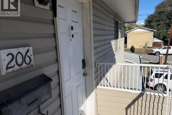 Exterior view of a white door and windows with grey siding, number 206 sign, and a white railing. - 3712 24 Avenue Unit# 206, Vernon