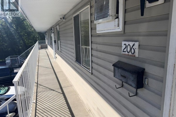 View down a long balcony with white railings, grey siding, and windows, leading to apartment door 206. - 3712 24 Avenue Unit# 206, Vernon