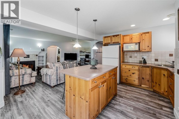 Lower level kitchen and living area with a fireplace, couches, and wood cabinetry on the kitchen side. - 3712 Cavendish Blvd, Nanaimo